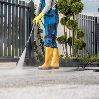 Cleaning the Driveway with Pressure Washer Man in Yellow Rain Boots and Work Uniform Pressure Washing Concrete Tiles of the Driveway to His House Behind the Closed Yard Entrance Gate. Home Surroundings Maintenance Theme.